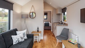 Un espacio habitable pequeño con sofá, una mesita con una botella y vasos, una silla, una zona de cocina con fregadero y armarios, y un ventanal con vistas a zonas verdes.