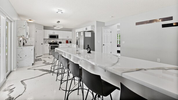 A modern kitchen with a long island counter, bar stools, and white cabinetry. - Coastal Retreat - Roomy, Relaxing Heated Pool (Palm Coast)