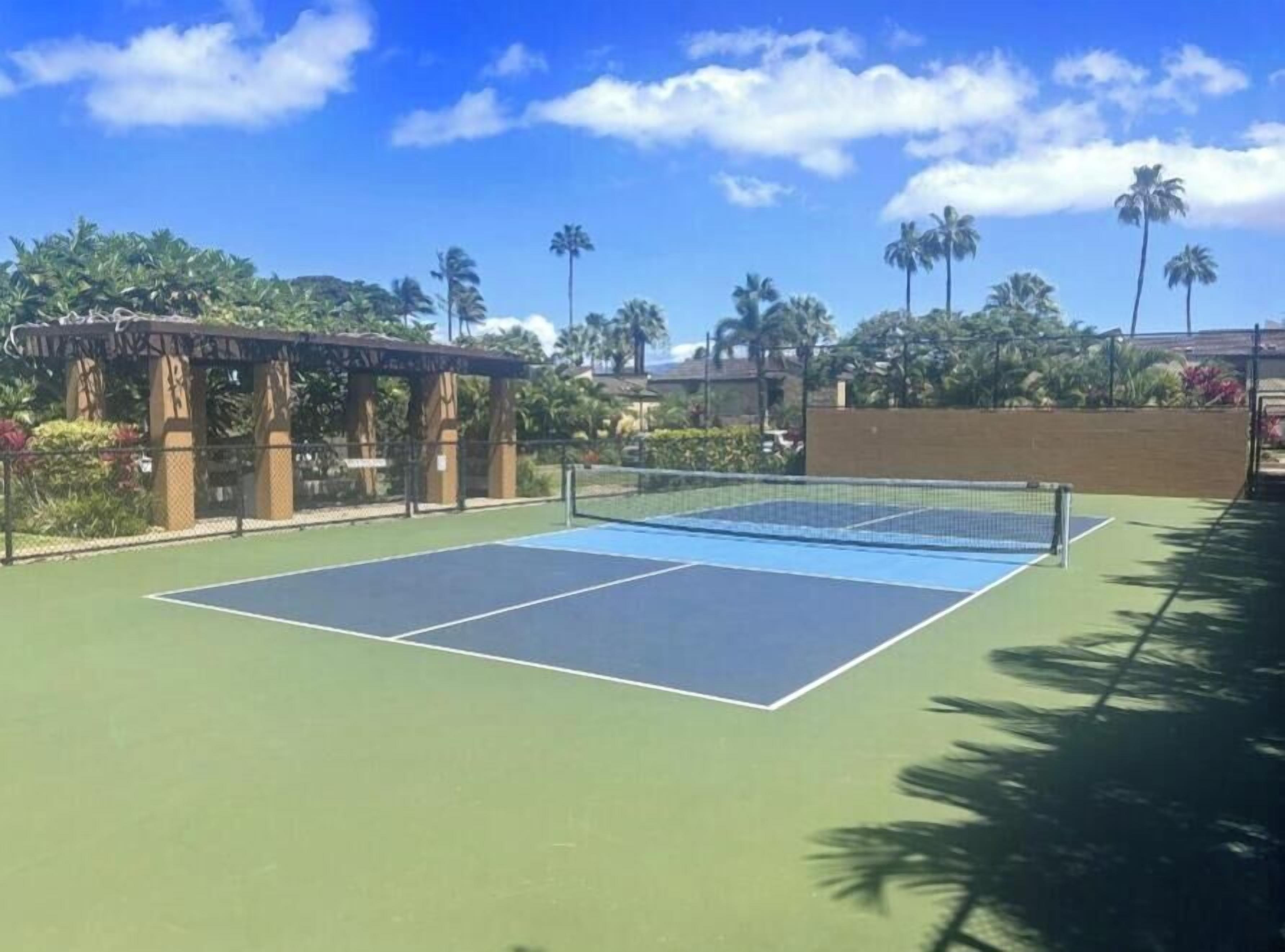 An outdoor tennis court with a net, surrounded by a wooden pergola and palm trees.