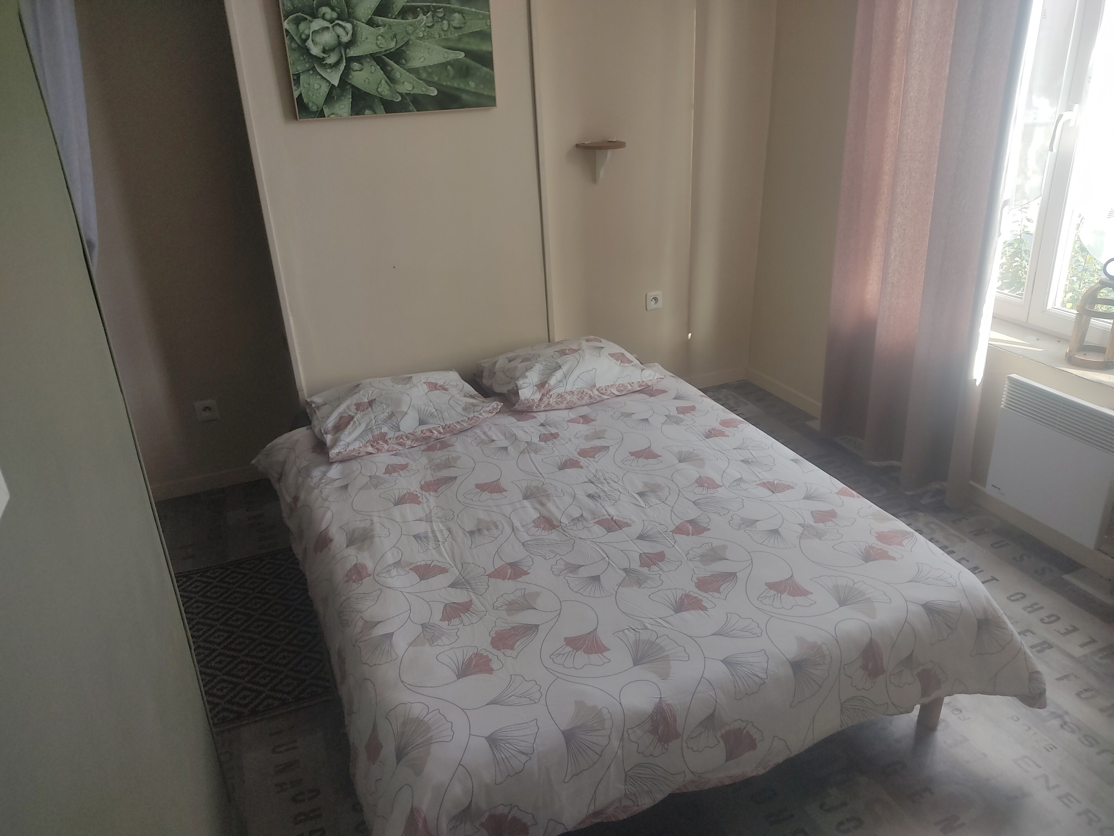 A small, neatly made bed with floral bedding, a wall-mounted light switch, and a window with curtains.