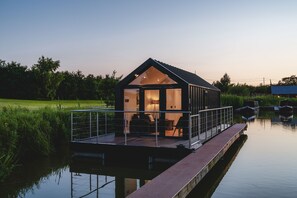 A modern houseboat with a glass facade, a deck, and a small outdoor seating area. - Whimbrel (Chester)