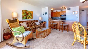 A living room with a sofa, chairs, a glass coffee table, and a kitchen area with wooden cabinets and a microwave.