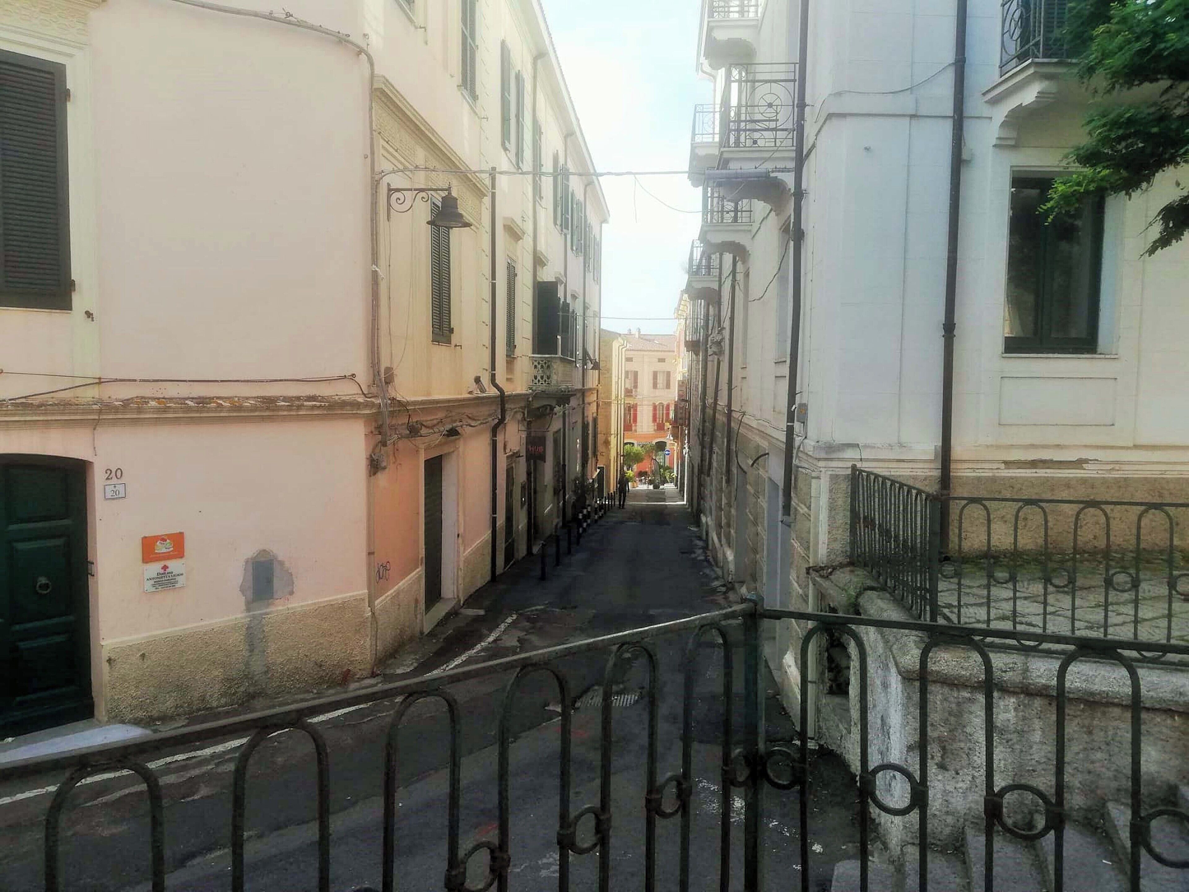 Une rue étroite bordée d’immeubles dotés de balcons, avec une clôture en métal au premier plan.
