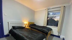 A room with two beds, a window with curtains, a radiator, and a wall socket. - 3 Room Apartment - Twins-double (King's Lynn)