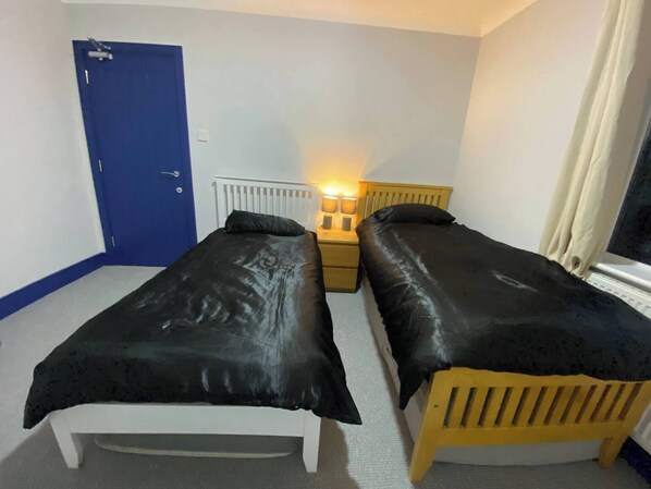 Quarto com duas camas de solteiro, uma porta azul, mesa de cabeceira com abajur e janela com cortinas.