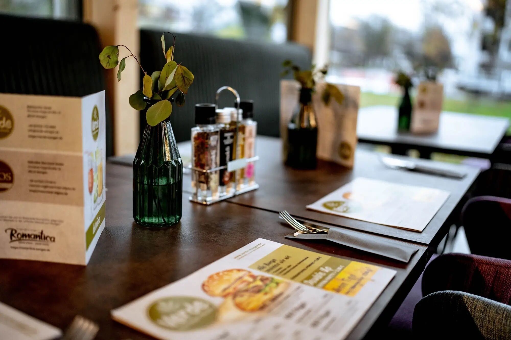 Una mesa de restaurante con un menú, una botella verde con una planta y condimentos.