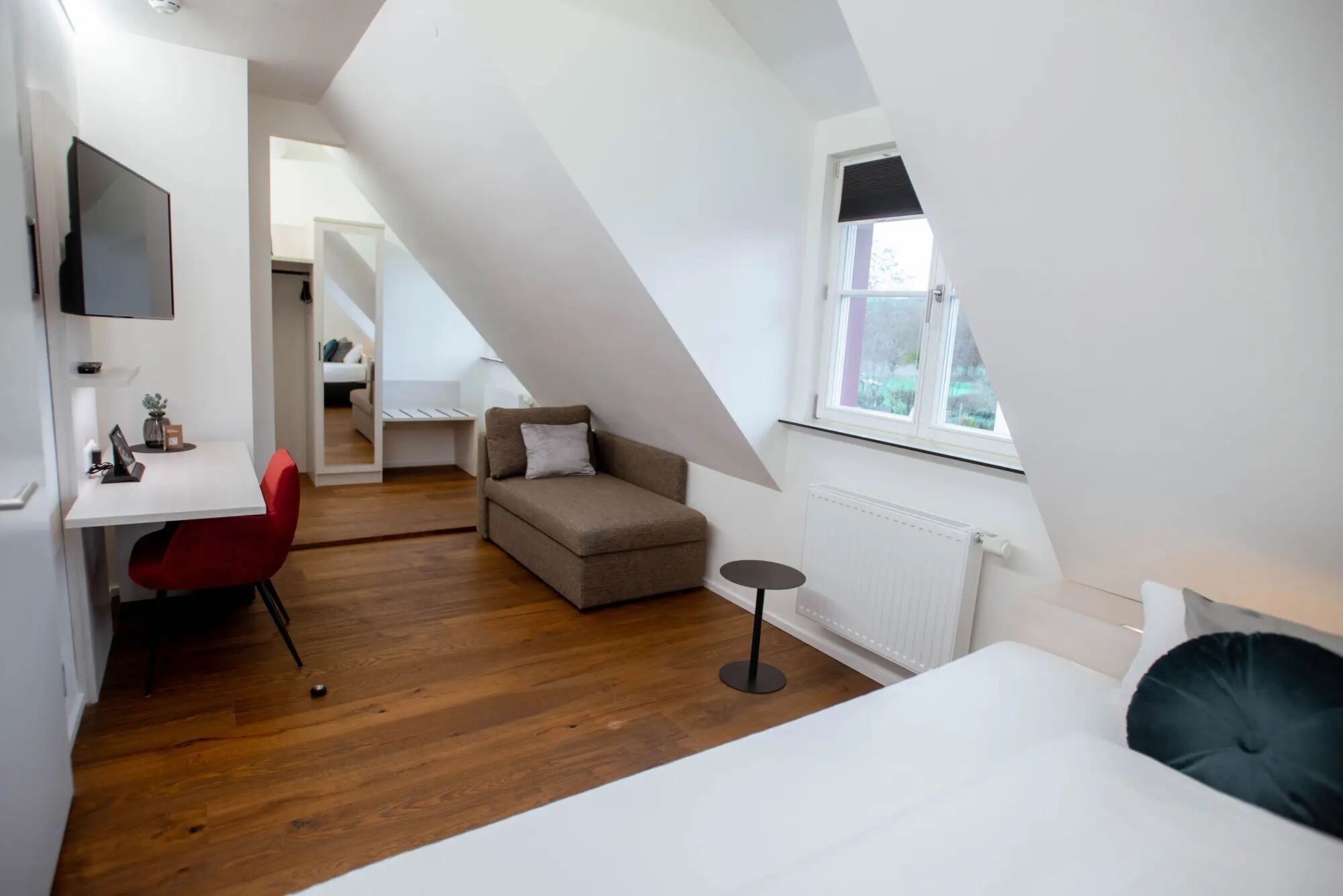 Une pièce moderne et bien éclairée, avec un plancher en bois, un lit, un bureau avec une chaise, un canapé et un téléviseur fixé au mur.
