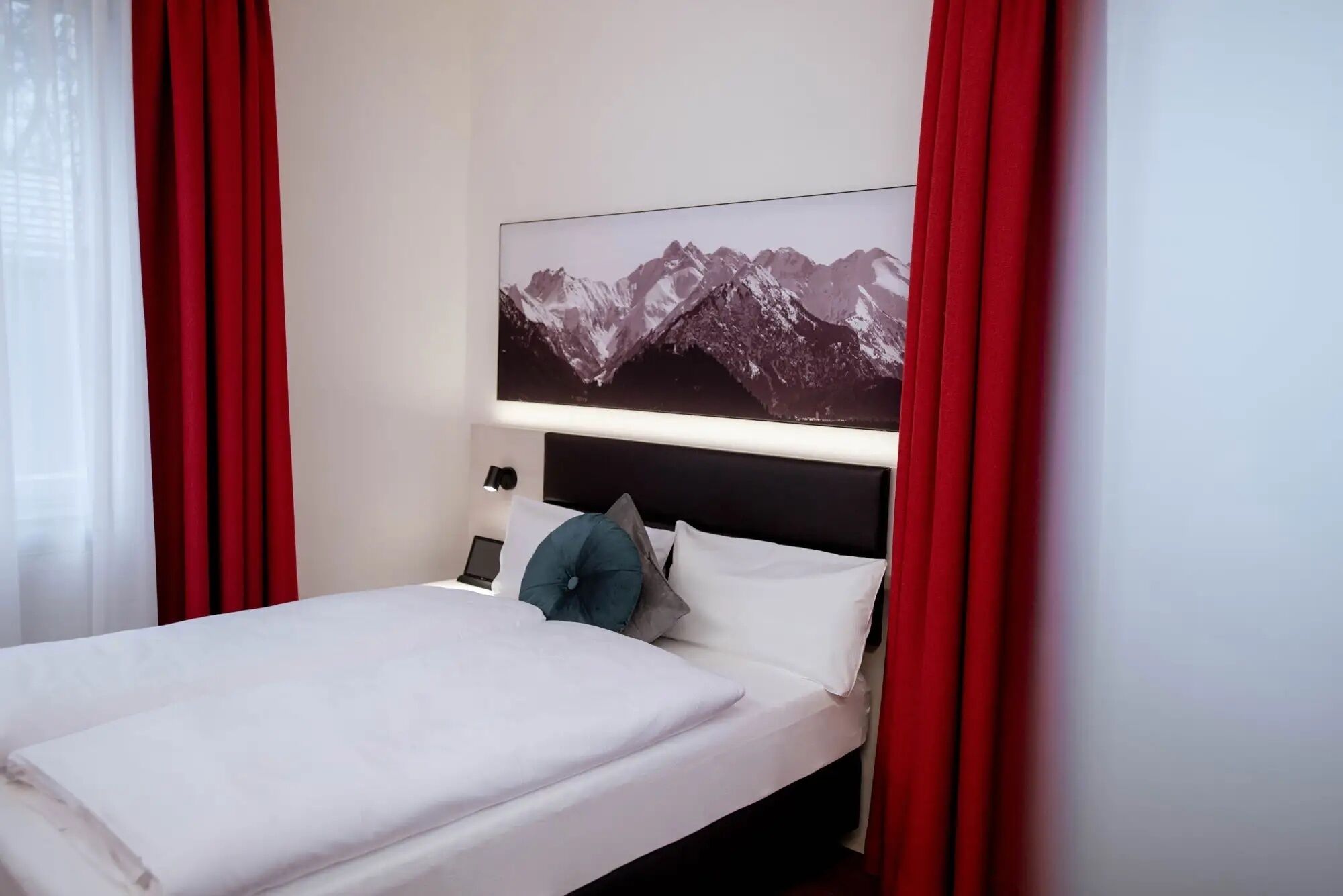 A hotel room with a bed, a nightstand, a lamp, and a mountain landscape painting on the wall.