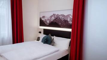 A hotel room with a bed, a nightstand, a lamp, and a mountain landscape painting on the wall.