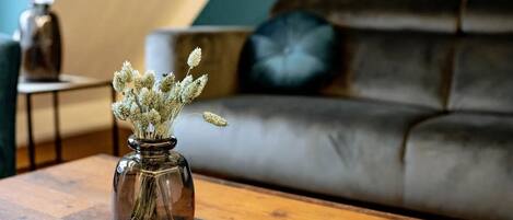 Un salón con un sofá de cuero marrón oscuro, una mesa de centro redonda de madera y un jarrón de cristal transparente con plantas secas sobre un posavasos gris.