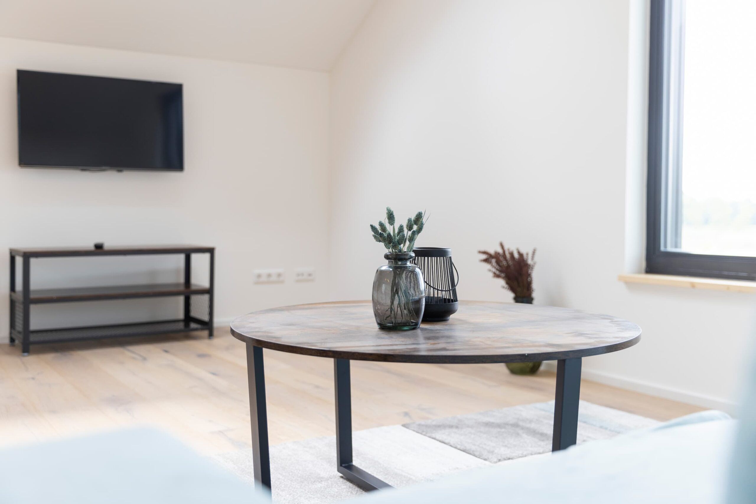 Una sala de estar moderna con una mesa redonda, un televisor de pantalla plana montado en la pared y un ventanal que proporciona luz natural.