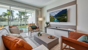 A modern living room with a sofa, coffee table, and chairs, a flat-screen TV mounted on the wall, and a balcony with a view of greenery. - Spacious Modern new Home With Private Pool (Kissimmee)