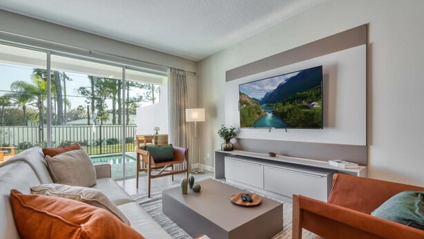 A modern living room with a large flat-screen TV, a sofa, a coffee table, and a view of a pool and greenery outside. - Luxury new Home Close to the Parks (Kissimmee)
