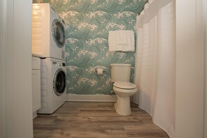 A compact bathroom with a washing machine, toilet, and patterned wallpaper. - A Wave From It All - Studio 10 Min to Ocean (Myrtle Beach)