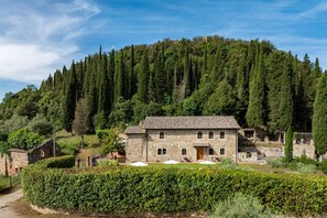 A stone building with a tiled roof, surrounded by greenery and a forested hill. - Podere Montioni 6+2, Emma Villas (Rapolano Terme)