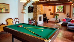 Une table de billard avec des billes, un canapé, un piano et une cheminée dans une pièce aux murs en pierre et aux poutres apparentes.