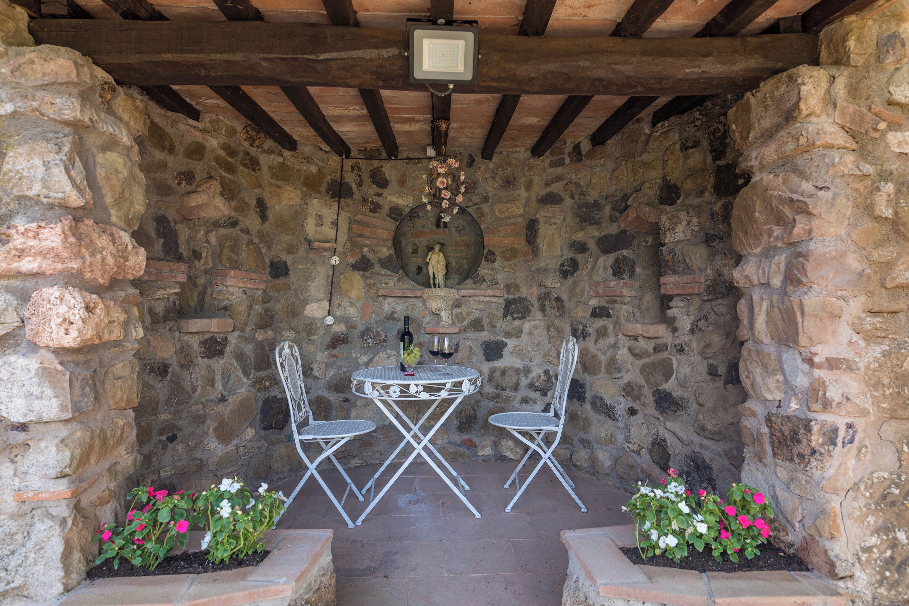 A stone-walled outdoor seating area with a table and chairs, a bell, and a small statue.
