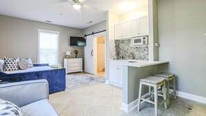 Un soggiorno moderno con un divano blu, una piccola zona cucina con mobili bianchi e un forno a microonde, e una ventola a soffitto.