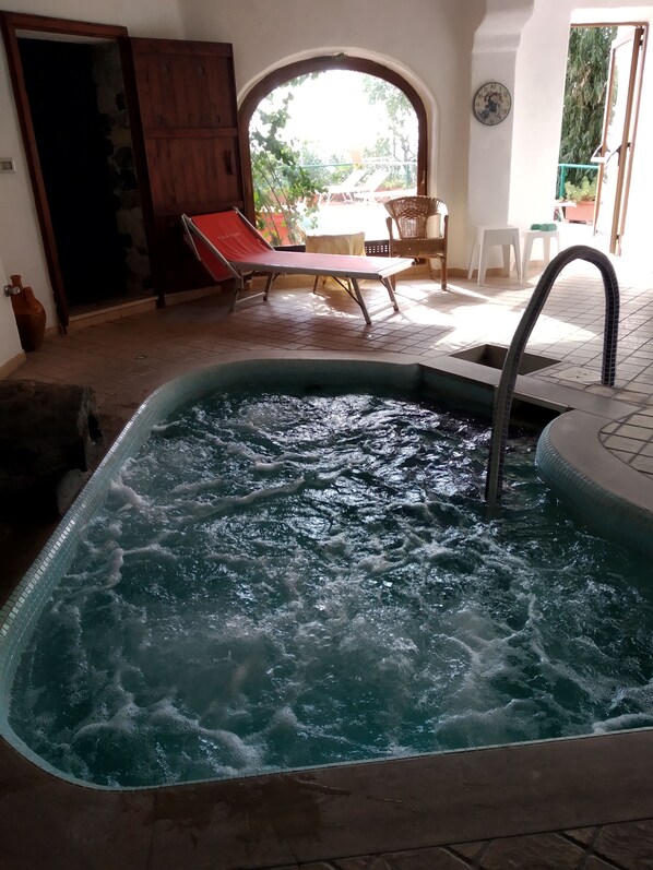 A hot tub with a tiled interior, a wooden door, and a view of an outdoor seating area with a table and chairs. - Hotel Villa al Mare (Barano d'Ischia)