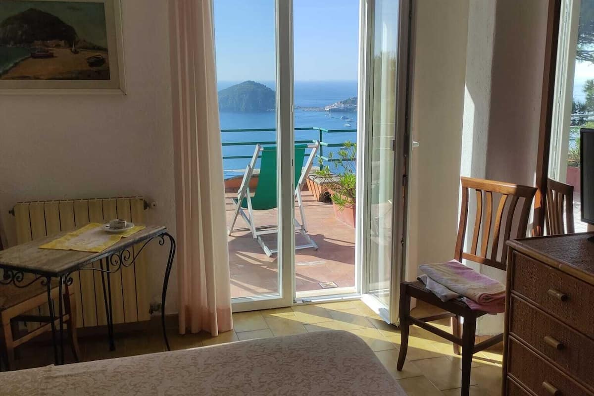 Quarto com cama, escrivaninha, cadeira e cómoda. Há uma porta de correr para área externa com vista para o mar e uma montanha.