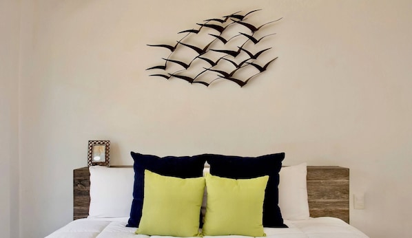 Un dormitorio con una cama, dos almohadas, una obra de arte en la pared y una mesita de noche con un cuadro enmarcado.