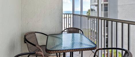 A balcony with wicker chairs and a glass table, overlooking a beach and a building with balconies.