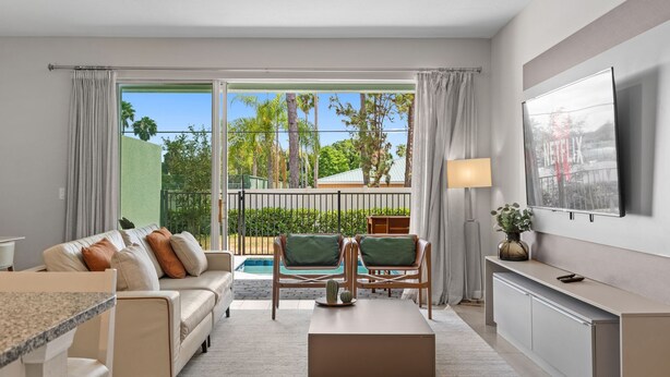 A modern living room with a sofa, coffee table, and chairs, a TV mounted on the wall, and a view of a pool and greenery outside.