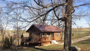 A wooden cabin with a porch, surrounded by trees and a grassy field.