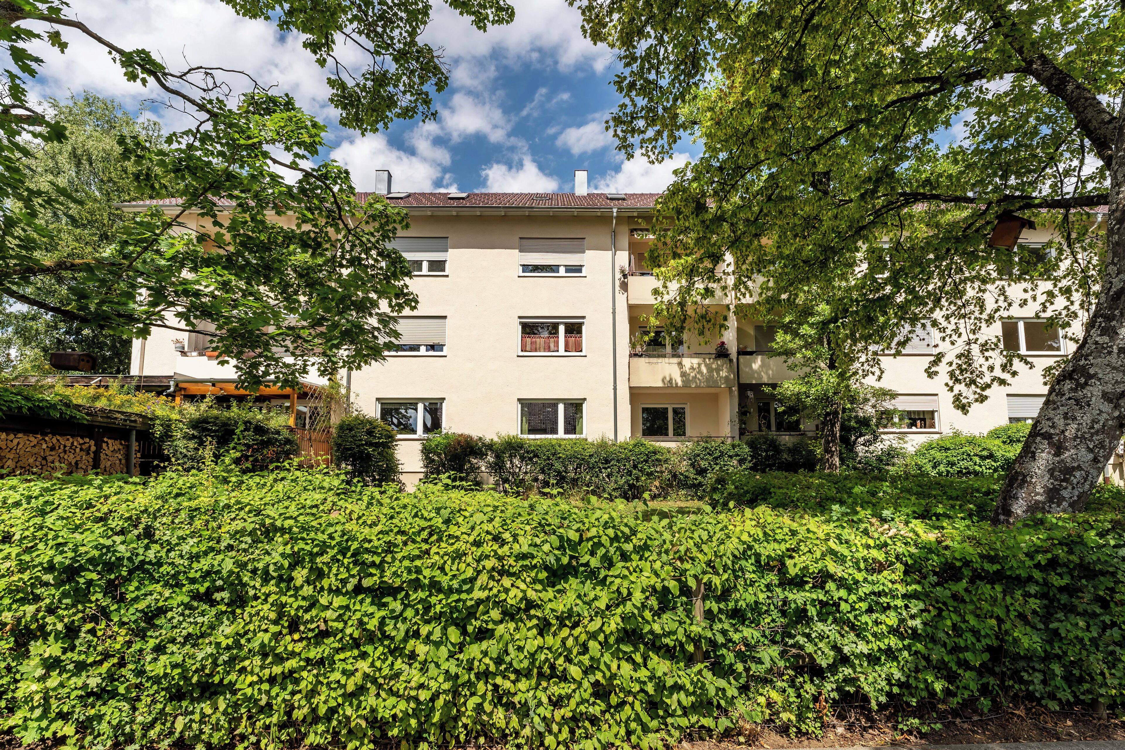 Ein mehrgeschossiges Wohngebäude mit hellbeiger Fassade, umgeben von üppiger Bepflanzung und Bäumen.