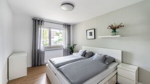 A modern bedroom with a bed, bedside tables, a window with curtains, a radiator, and a small white cabinet. - City-Luxury Apartment Rottweil (Rottweil)