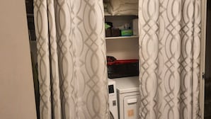 Un baño con cortina de ducha, lavadora y repisas con artículos de aseo.