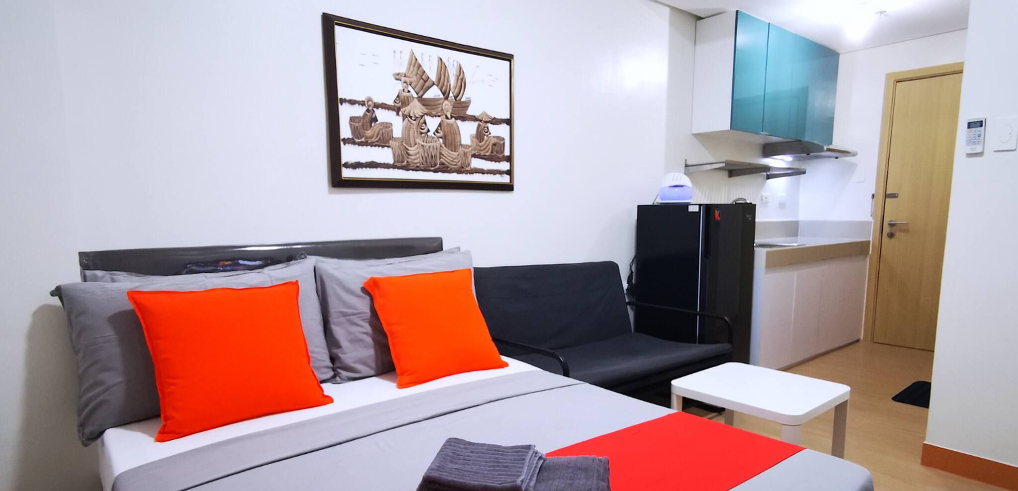 A hotel room with a bed, two orange pillows, a black sofa, a small white table, and a kitchen area with a stove and microwave.