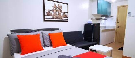 A hotel room with a bed, two orange pillows, a black sofa, a small white table, and a kitchen area with a stove and microwave.
