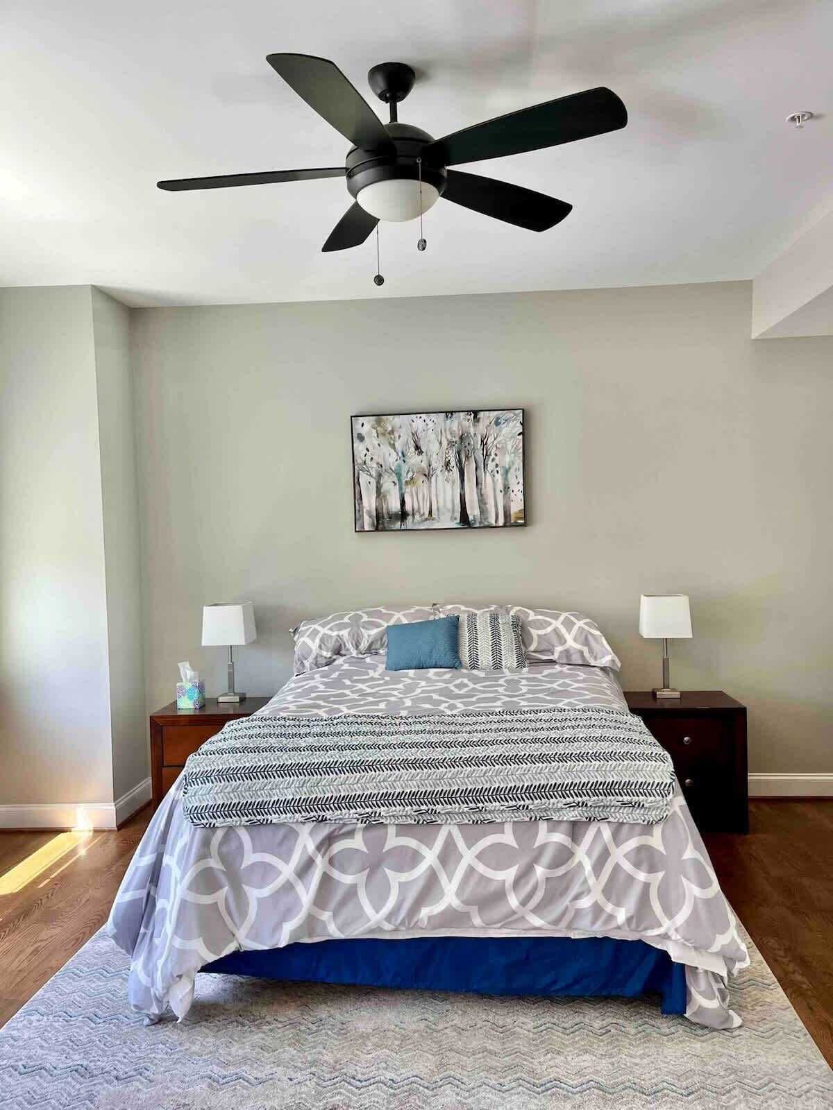 A bedroom with a bed, two bedside tables, a ceiling fan, a wall-mounted painting, and a patterned rug.