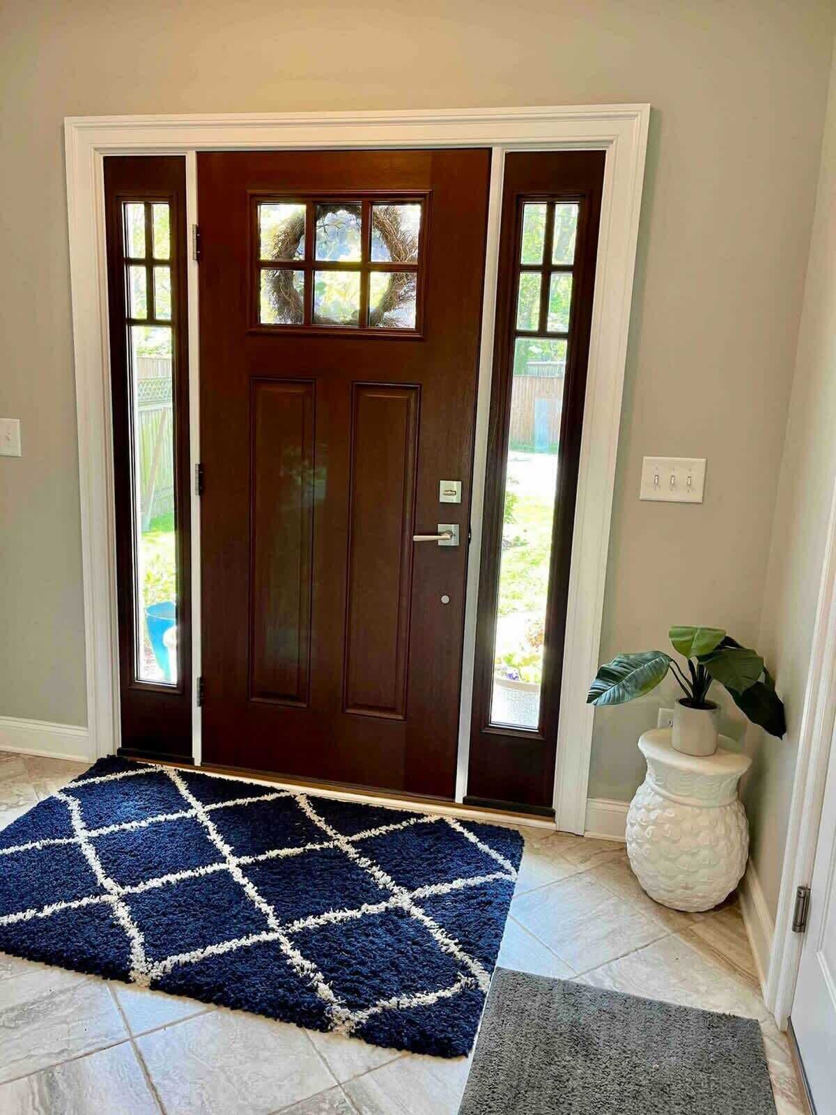 A dark wooden front door with a glass panel, a blue and white patterned rug, and a white textured planter with a plant.