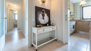 A hallway with a white console table, a wall-mounted light fixture, and a kitchen area with cabinets and a window.
