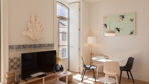 Una sala de estar moderna con un televisor de pantalla plana, una mesa de comedor con sillas, un elemento decorativo en la pared y un ventanal con vistas a edificios.