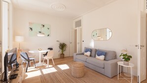 Una sala moderna con un sofá gris, una mesa redonda de centro y un televisor. Hay un comedor con sillas y una mesa, además de una planta en maceta.