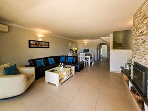 Amplia sala de estar con sofá, mesa de centro y mesa de comedor. Se observa una chimenea a la derecha, y una escalera conduce a un nivel superior.
