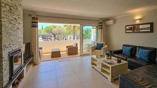 A living room with a fireplace, a sofa, a coffee table, and a balcony with a view of trees and a building. - FLH Quinta das Salinas Triplex with Terrace & Pool (Vale do Lobo)
