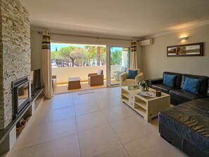 Een woonkamer met een open haard, een bank, een salontafel en een balkon met uitzicht op bomen en een gebouw.