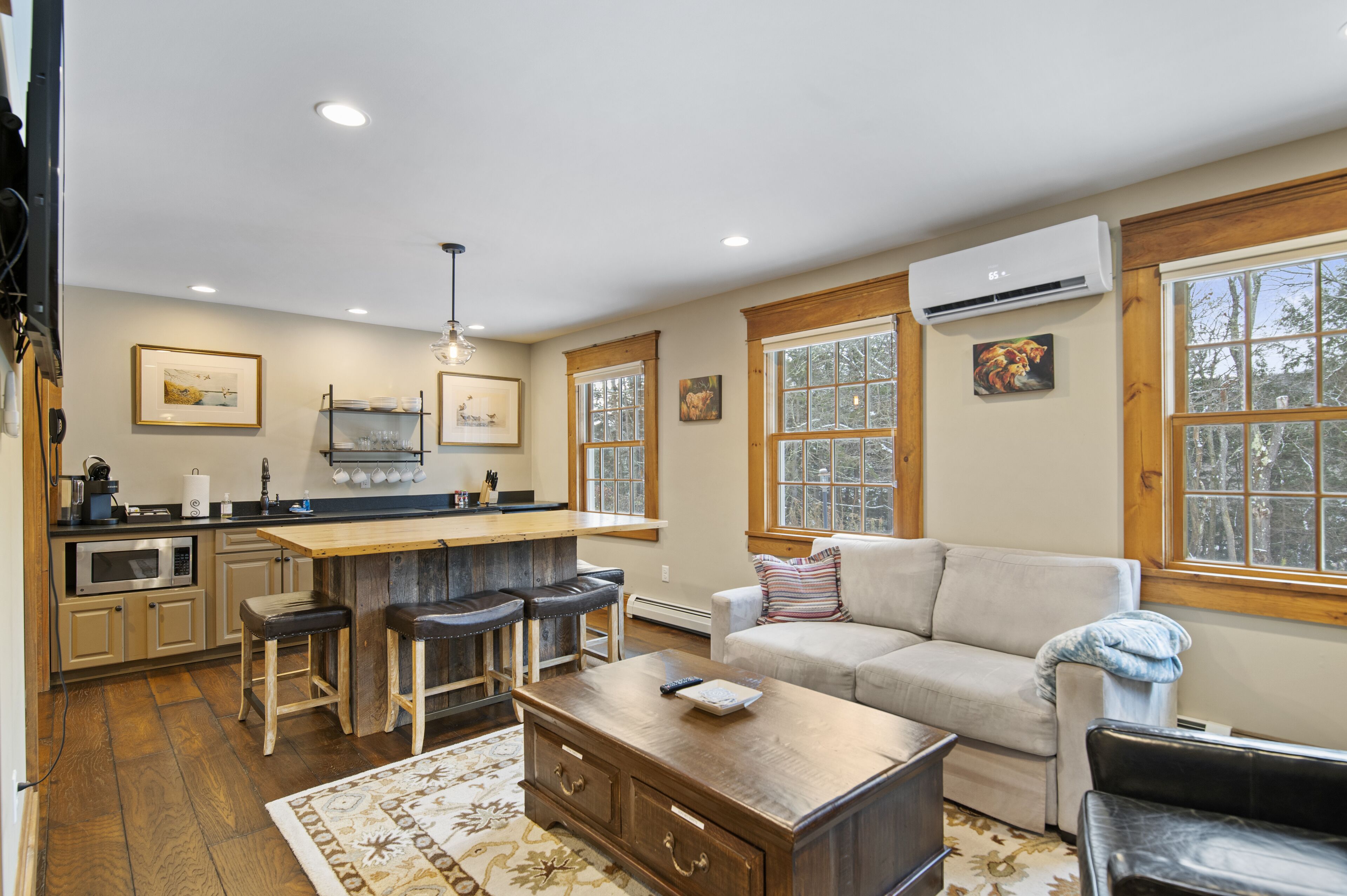 A modern kitchen with a central island, bar stools, and a dining table. There is a living area with a sofa and a coffee table.