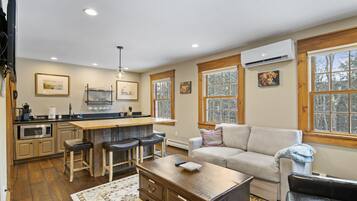 A modern kitchen with a central island, bar stools, and a dining table. There is a living area with a sofa and a coffee table.
