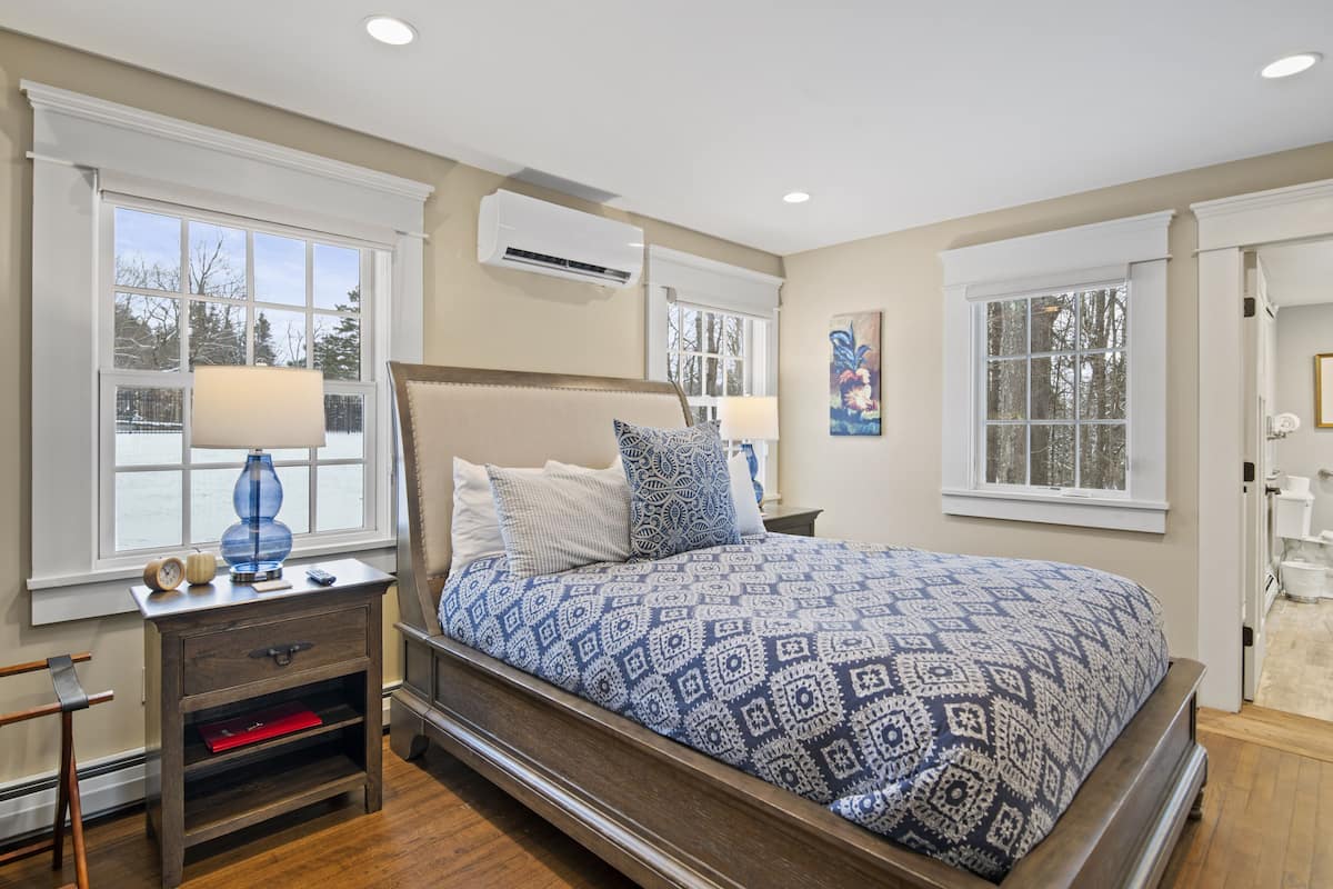 A bedroom with a large bed, two bedside tables, a nightstand, a window with a view, and an air conditioning unit.