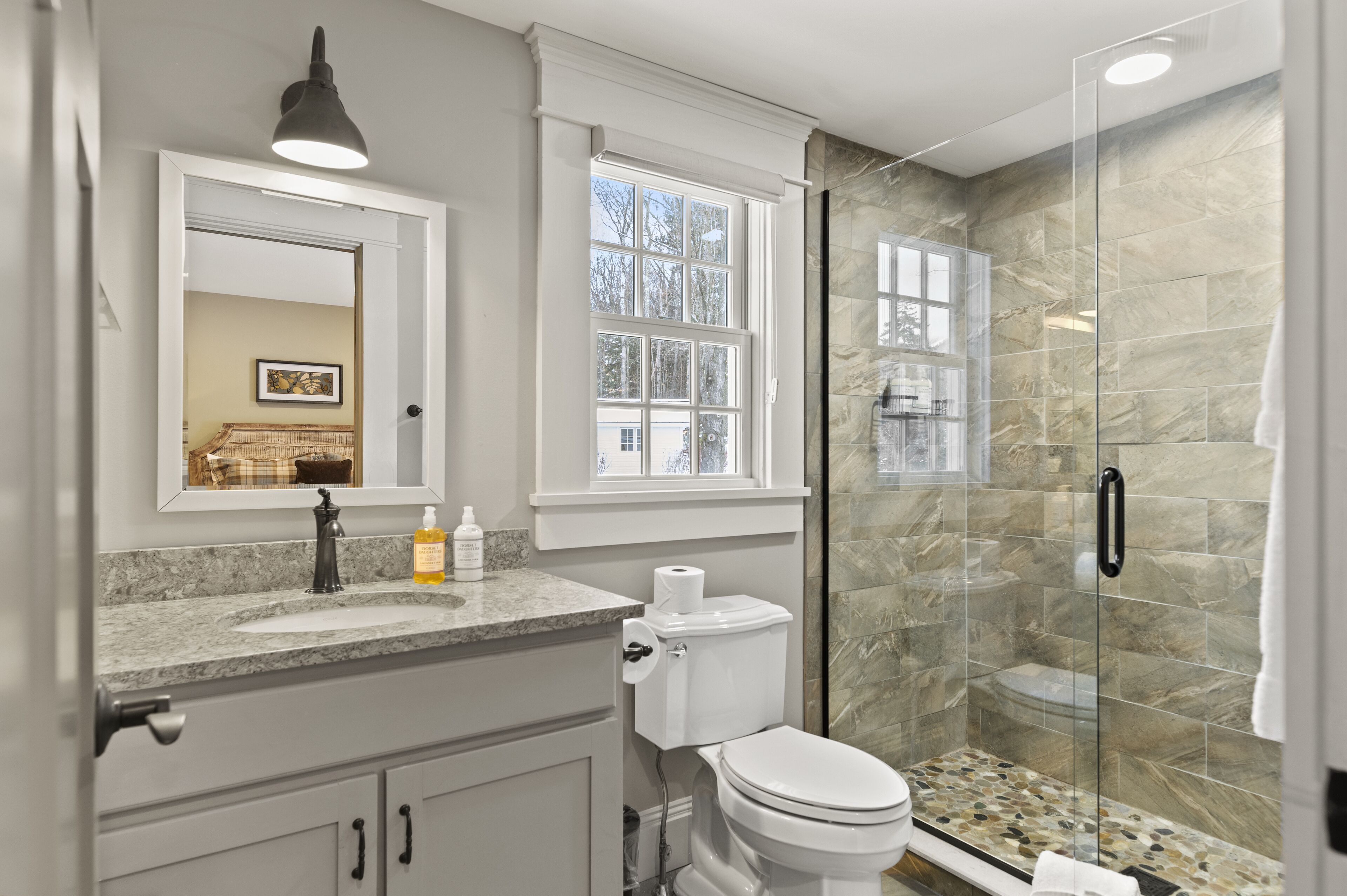 A bathroom with a granite countertop, a white toilet, a glass-enclosed shower, and a window above the toilet.