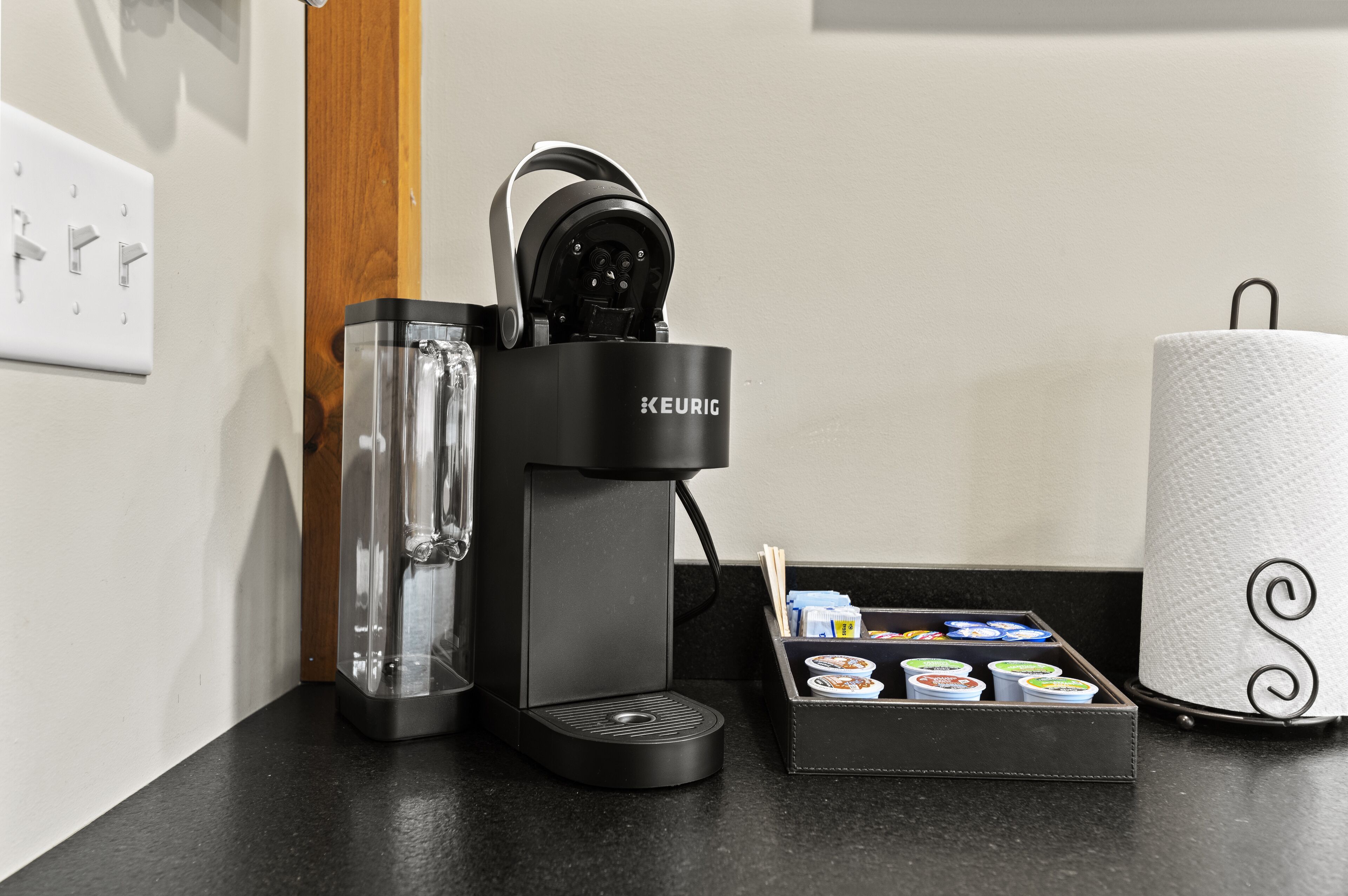 Una cafetera Keurig, una jarra de vidrio, una caja de cápsulas de café y un rollo de papel higiénico sobre una encimera negra.