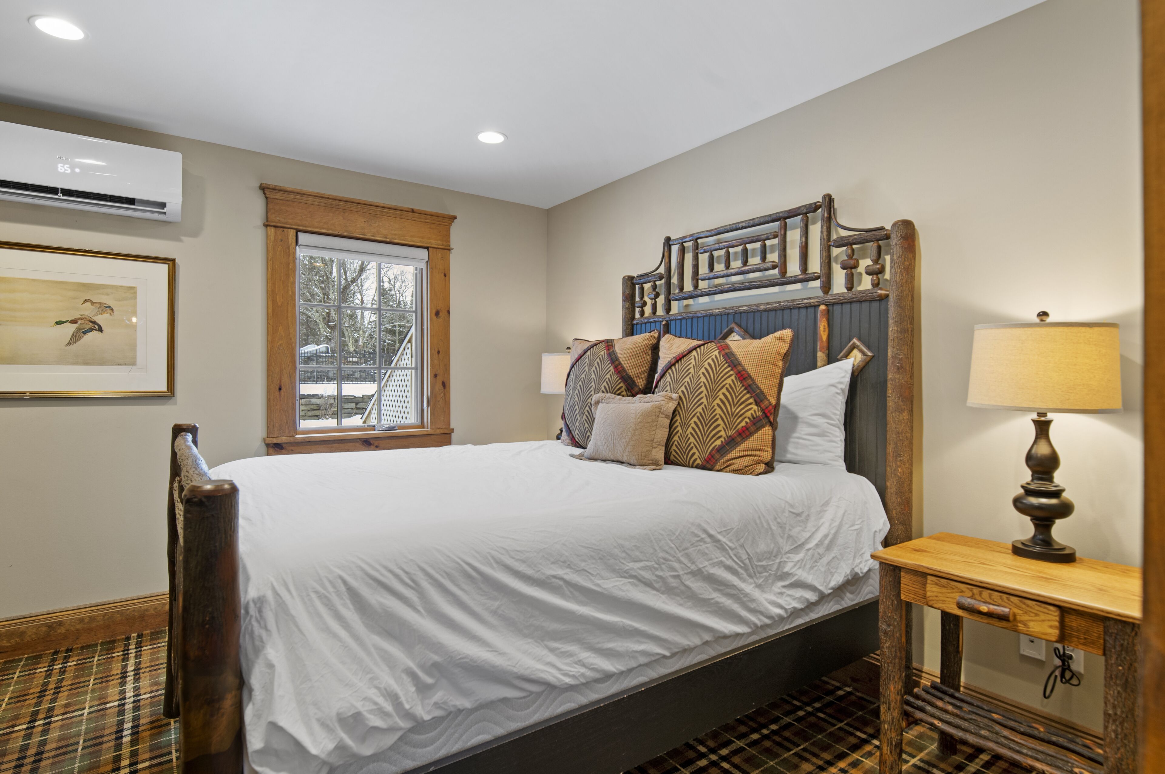 A bedroom with a large bed, wooden headboard, a bedside table with a lamp, a framed picture on the wall, and a window with a view of trees.
