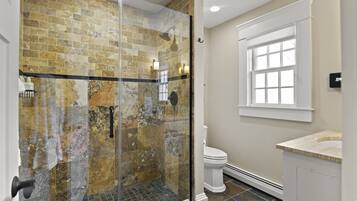 Une salle de bain avec une douche à parois de verre, des toilettes blanches, un lavabo beige et une fenêtre aux montants blancs.
