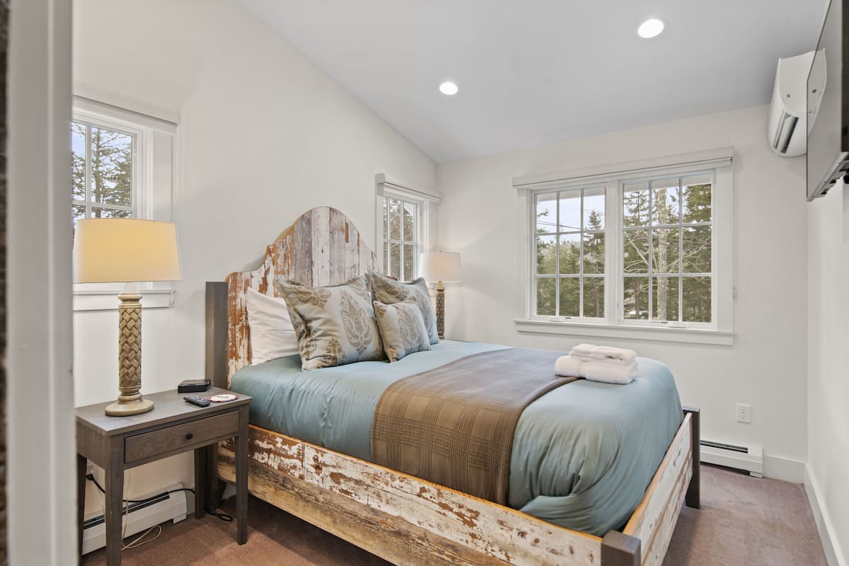 A bedroom with a rustic wooden headboard, a bed with multiple pillows, a bedside table with a lamp, and a window providing natural light.