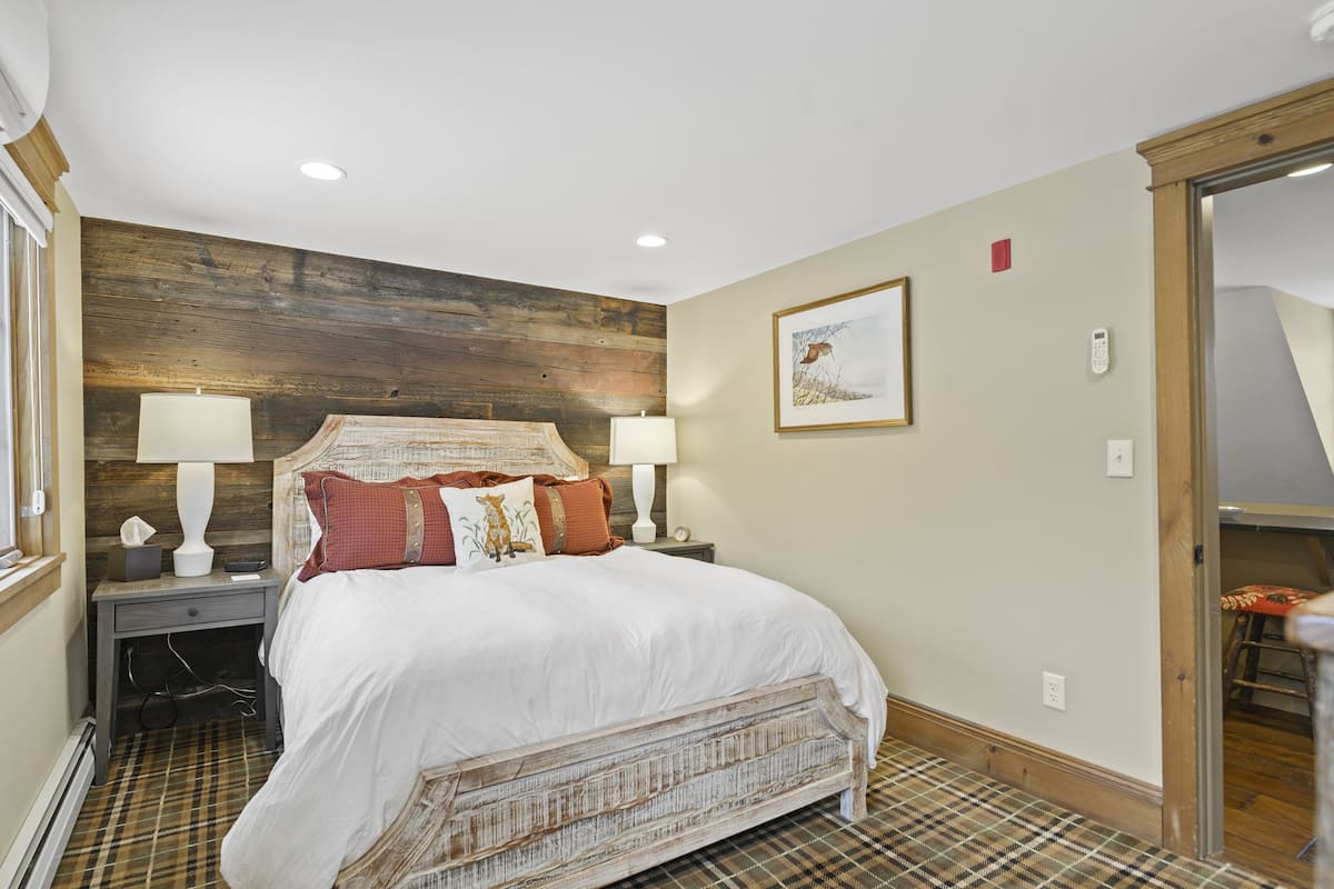 A bedroom with a wooden headboard, a large bed with white bedding, two bedside tables with lamps, and a plaid-patterned floor.